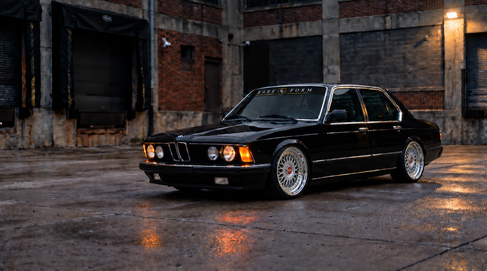 Rendering of Shawn’s black 1985 BMW 735i with a RAREFORM windshield banner, on BBS RC090 wheels, outside a brick warehouse at dusk